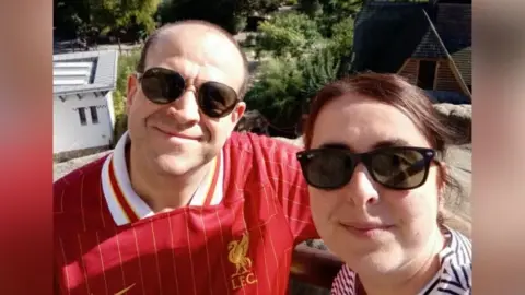 Allister Cubitt Two people take a sunny outdoor selfie in front of a number of buildings and greenery. The bald man on the left wears a red Liverpool FC jersey and sunglasses, and the brown-haired woman on the right wears a black‑and‑white striped shirt and sunglasses.