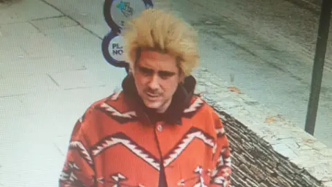 Alexander Key is walking in a street. He has long peroxided hair standing up and a moustache. He is wearing a bright red coat which has a bold zigzag print in white and black.