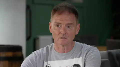 A head and shoulder shot of Mark Terry-Lush looking into the middle distance. He's wearing a grey top. Behind him the background is blurred but there's a barrel next to his left shoulder and chairs next to his right.
