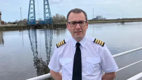 Captain Paul Brookes is standing near the River Tees, with white metal railings behind him. Beyond the river are the blue legs of the Middlesbrough Transporter Bridge, also reflected in the water. Brookes has short, dark hair and black glasses, is clean shaven, and is wearing a white, short-sleeved shirt with black epaulettes with four gold bands on each. 