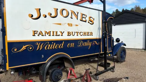James Miller A pre-war blue van, with yellow livery around its doors, windows and edges. A sign painted on the side of the rear of the van says "J.JONES, FAMILY BUTCHER, Waiting on Daily". The rear right wheel has been removed and various tools are on the ground beside it.