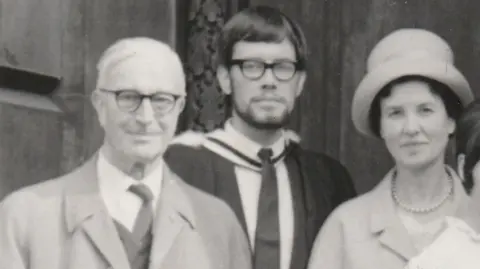 John Harrison A black and white photograph of John Harrison pictured in 1965. He is dressed in a suit and graduate gown and has dark hair and dark framed glasses. A man and a woman are stood either side of him. The man is wearing a suit and coat and dark framed glasses. The woman is wearing a light coloured coat and hat.