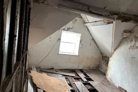 Inside the chapel. There are wooden floorboards in disarray, and the room has a pitched roof with a window. The white plaster on the walls has flaked off in areas and wooden beams are visible on the left-hand wall.