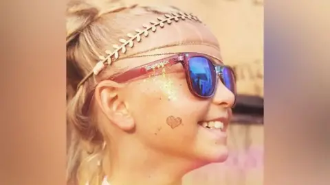 Mason Family A close-up of Kezia who has blonde hair styled in braids, wearing a decorative leaf-patterned headband. Warm lighting gives the image a soft, golden tone.