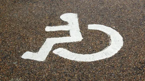 A white accessibility symbol on an asphalt surface. It is the familiar icon of a person seated in a wheelchair, typically used to mark disabled parking bays or accessible routes. 