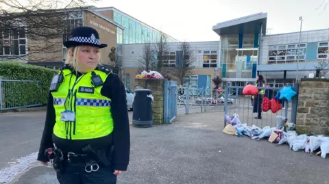 A police officer in a hi-vis vest outside the school gates. Numerous floral tributes have been placed along the right side of the gate and wall, together with balloons.