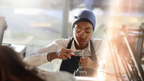 A T-level jewellery student working at a bench making an item of jewellery. She has a headscarf around her neck and holds tools in her hands. She wears a white top with a black apron over it and is looking down at what she is working on.