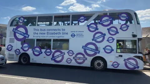 Double decker bus outside Abbey Wood station. There is a banner on the side that reads, "This bus connects to the new Elizabeth line". It is white and covered in images of Elizabeth line station roundels.