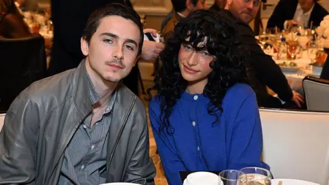 Getty Images Odessa poses with Timothee Chalamet for a picture sitting next to each other on a table. Odessa is in a blue jumper whilst Timothee is a wearing a grey stripy shirt and a grey leather jacket. 