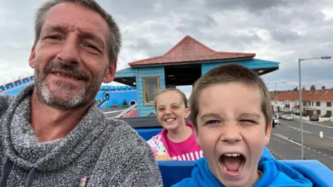 Coleman family Jason Coleman, Freddie Coleman and a young girl sitting on what appears to be a rollercoaster at a theme park. Jason is wearing a grey and white hoodie, has short grey hair and is smiling. Freddie has short brown hair, is wearing a blue hoodie and has his mouth open in excitement. The young girl is smiling while wearing a pink T-shirt. She has light coloured hair which has partially blown across her face.