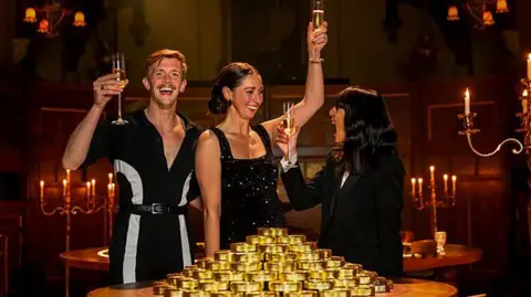 BBC/Studio Lambert/Paul Chappells Stephen holding a glass of champagne standing next to two women. One of the women, Rachel is wearing a black sparkly dress with her dark hair tied back. The other is Claudia Winkleman, who is also wearing black. The are both also holding champagne glasses. In front of the is a pile of gold coins. They are standing in a grand old-fashioned room surrounded by candelabras.