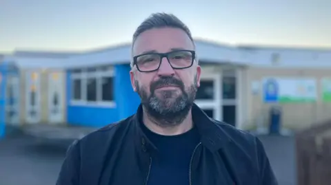 BBC / Elizabeth Baines Nicholas Humphrey looks at the camera. He is wearing black glasses, navy top and black coat. The school is in the background.