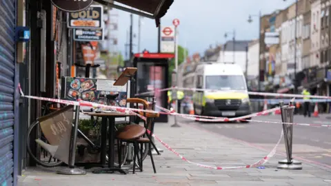 PA Media The exterior of a restaurant on a London street, the tape from a police cordon is wound around the crime scene, a chair is slightly tipped up beside a table. 