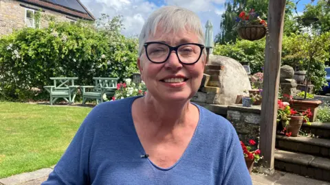 A woman sits in the garden of her home. She is wearing a blue top and has grey hair and dark glasses. There are plants in pots in the background. Another house is to the left of the image. There is also a hedge in the background.