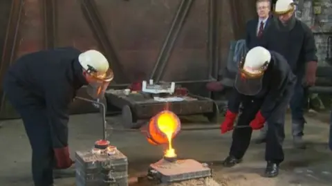BBC Two men in protective clothing pour molten metal into a bell cast