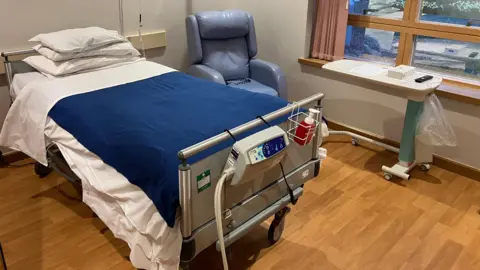 A room at Loros Hospice with a bed and chair next to a window.