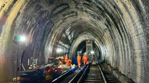 The Blackheath tunnel inspected by four men in orange jackets plus another using ladder. Equipment lays on the tracks to the left.