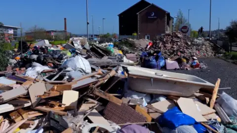 Glastonbury Gabriel A huge site with rubbish strewn all across it. There's a bath in the foreground, lots of large planks of wood and card and a pile of bricks in the background in front of a Premier Inn building. The sky is blue