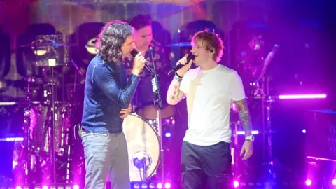 Dave J Hogan/Getty Images Gary Lightbody of Snow Patrol and Ed Sheeran singing together onstage in a purple-lit set at Latitude Festival. Lightbody who has shoulder-length dark hair, is wearing light grey trousers and a blue long-sleeved top. Sheeran is wearing navy blue trousers and a white T-shirt. A drummer is playing behind them.