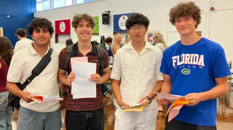 Four teenage boys stood in a row. They are all holding sheets of paper, smiling and looking at the camera.