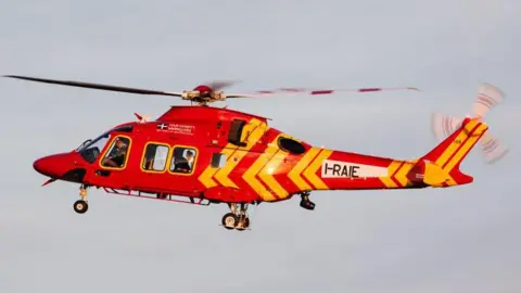 A red helicopter with yellow markings is flying and part of the rotor appear blurred because of the movement.