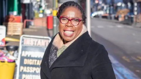 A woman with black hair, red glasses, a scarf and black coat, stands smiling at the side of a road.