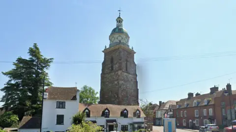 A white building with a thatched roof sits adjacent to a large brick tower. A dome structure sits at the top of the building, alongside a cross. Rows of houses can be seen surrounding the building.
