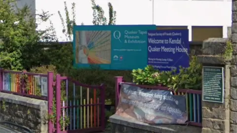 Google The Quaker Tapestry Museum in Kendal. It is an old Georgian house with two signs in front advertising the museum and a sign reading 'welcome to Quaker meeting house'. The entrance to the car park has coloured railings.