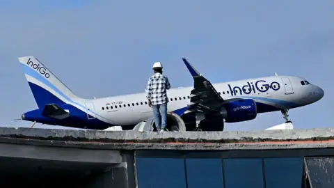 AFP via Getty Images A man stands atop an under-construction building as an Indigo aircraft prepares to land at Kempegowda International Airport in Bengaluru 