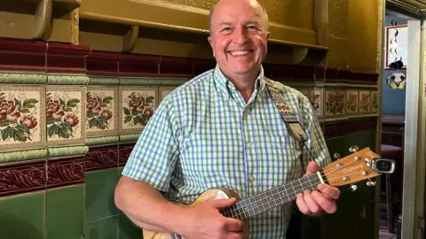A man is wearing a blue and green gingham shirt and is holding a ukulele that is swung over his shoulder. He is smiling and standing in front of green and red tiles.