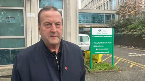Phil Bialyk wearing a black zip neck top and a navy blue jacket and a poppy badge and to his right there is an Exeter City Council Civic Centre sign in green and white.