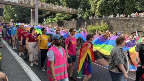 Crowds taking part in the Pride march in 2024. People are walking in the road and are holding a long rainbow coloured flag.
