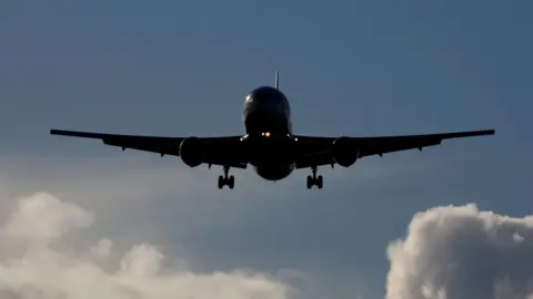A Boeing 767 aircraft is silhouetted against a blue sky with fluffy white clouds. The aircraft is coming towards the camera with its wheels down ready to land.