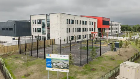 A modern three-storey school building with light brick walls and a red entrance feature stands behind a fenced outdoor sports court. Newly planted trees, landscaped grounds, and pathways surround the campus, while a project sign sits in the foreground under an overcast sky.