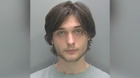 A custody photo of Jonathon Austin wearing a grey top. He is staring but not smiling. He has blue eyes, facial hair and long brown hair.
