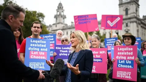 Getty Images Kim Leadbeater, wearing a navy jacket and blue top, being interviewed by a journalist with a microphone. Behind her are a number of campaigners holding placards in support of the assisted dying bill. Their signs say "Legalise assisted dying and let us choose".