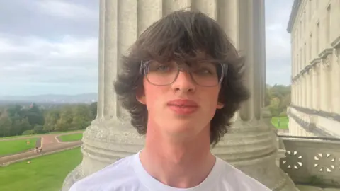 BBC Darragh wears glasses and a white t-shirt. He has long dark hair.