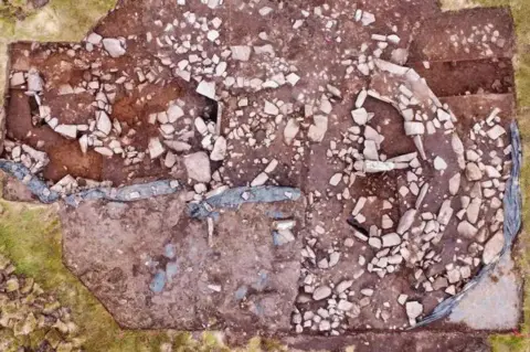 Tris Thorne The aerial view of the prehistoric structure at Spurness shows large stones laid out on an area of red soil. Some of the stones have been laid out in such a way they look like walls and chambers. 