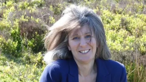 Supplied Heather Wainwright outdoors with her hair blowing in the wind. She has blonde/white hair in a bob, blue eyes, and is wearing a blue jacket. She is smiling at the camera and behind her are some green bushes.