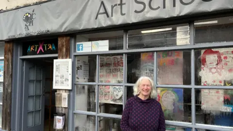 Katrina Atkinson is standing in front of her art school. She is wearing a blue jumper and has white hair. Art is hanging in the window of her shop.