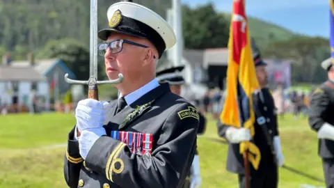 Andrew Cunningham carrying the Sword of State. He is wearing a dark coastguard uniform with gold decoration and a white and black cap. He is wearing black-rimmed glasses and white gloves.