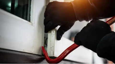Two gloved hands using a red crowbar to break open a window, which is painted white. 