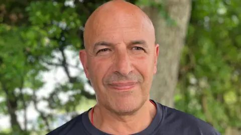 The image shows a person standing outdoors in front of trees and greenery, with the background blurred and out of focus. The photograph is a close-up portrait, with the person wearing a dark T-shirt and positioned against a wooded setting.