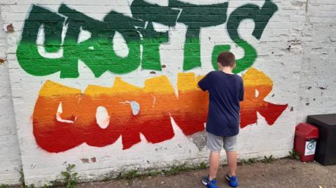A boy paints a Crofts Corner graffiti-style sign on the side of a building