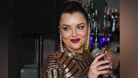 Roy's Shona sings inside the restaurant, with the bar behind her. She wears a gold and black sparkly long sleeved dress and her hair is tied back.