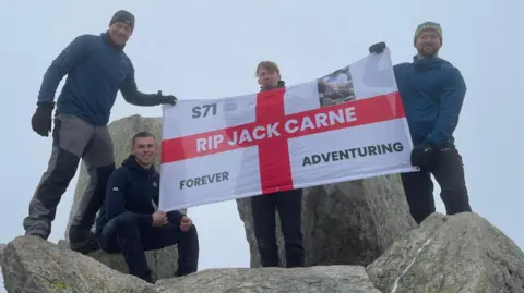 Matty Belcher | Brandan Smith Four men stand on top of large grey rocks holding up a st George's cross flag which reads 'RIP Jack Carne'