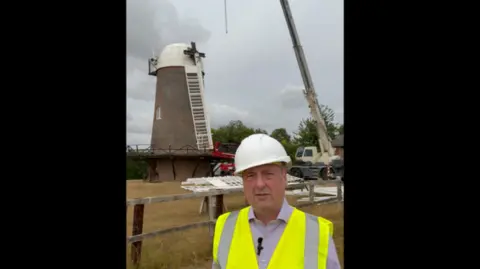 Wiltshire Council A brick windmill with a white rounded top and a large crane lifting a sail into place. A man in a high vis jacket stands in front of it.