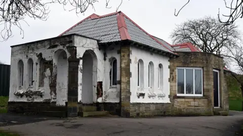 A small stone building with a white-painted entrance porch area. It is run down and seems to not be in use.