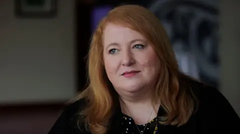 PA Media Naomi Long has long ginger hair and is wearing a black cardigan and dark patterned blouse 