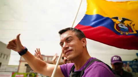 BBC Ecuadorian presidential candidate Daniel Noboa at a campaign event. He is giving a thumbs up to the crowd and holding a large Ecuadorian flag which billows over his left shoulder. He is also wearing a bullet-proof vest.